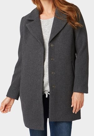 Donna che indossa un cappotto grigio scuro con bottoni sopra un maglione grigio chiaro e jeans blu, in piedi con le braccia rilassate ai lati.