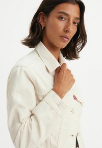 Chaqueta de mezclilla de color beige claro con un cuello clásico, botones frontales y detalles bordados en los puños. Presenta una etiqueta de logo roja en el bolsillo.
