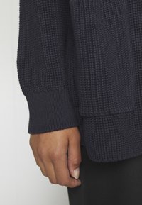 Granatowy sweter z dzianiny z teksturowanym splotem, ściągaczami na rękawach i luźnym krojem, podkreślający grubość i ciepło tkaniny.