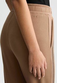 Hellbraune Jogginghosen mit elastischem Bund, seitlichen Taschen und dezentem Branding. Glatter Stoff mit tailliertem Design rund um die Hüfte.