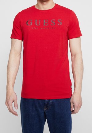 T-shirt en coton rouge avec un col rond. Présente le texte métallisé gris "GUESS LOS ANGELES" sur la poitrine. Manches courtes et coupe décontractée.