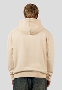 Beige hoodie with a drawstring hood, ribbed cuffs, and hem. Smooth cotton material, simplistic design, features a small logo patch at the waist.