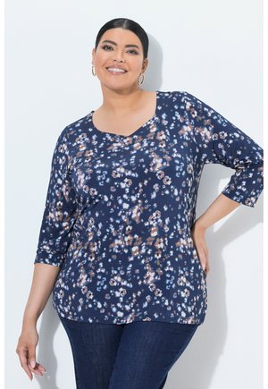 Smilende kvinde med mørkt hår iført store øreringe, en blå blomsterprintet langærmet top og mørke jeans, står med den ene hånd på hoften.