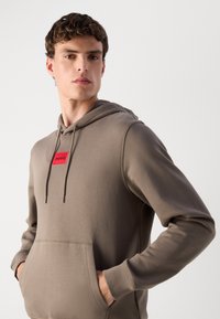 Felpa con cappuccio in verde oliva, realizzata in tessuto morbido. Presenta una toppa rettangolare rossa con il logo "HUGO", tasca a marsupio e lacci per il cappuccio.