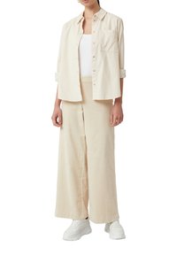 Camicia oversize beige chiaro abbinata a pantaloni larghi coordinati, con una tasca, e sneakers bianche. Tessuto morbido, vestibilità casual.