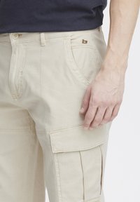 Beige cargo nadrág elöl zsebekkel és oldalsó zsebkifordítókkal. Aranyszínű logó díszítés a derékpánton. Simább, könnyű anyag.