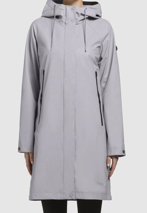 Femme portant un imperméable long à capuche gris clair avec fermeture éclair sur le devant et poches latérales zippées, debout devant un fond uni.
