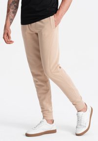 Beige jogger broek met zijzakken, voorzien van een gladde textuur en geribbelde enkelmanchetten, gedragen met witte sneakers en een zwart T-shirt.