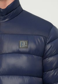 Tengerészkék pufi dzseki, magas gallérral, steppelt mintával és egy kis logófolttal a mellrészen, texturált, fényes kivitelben.