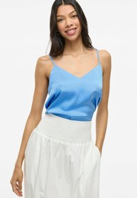 Svetlomodrý saténový top s V-výstrihom a tenkými popruhmi, skombinovaný s bielou sukňou so smockovaným pásom, vytvárajúci splývavý siluetu.