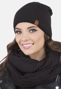Gorro de punto negro con textura acanalada, que presenta una pequeña etiqueta de cuero marrón. Combinado con una bufanda de punto grueso negra.