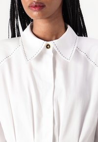 Camisa blanca de cuello con detalles de costura negra y un botón dorado en el cuello. Tejido suave con un diseño estructurado.