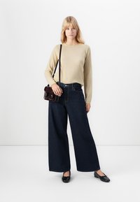 Kvinne iført beige langermet topp, mørke vide jeans, svarte leiligheter, og bærende en brun crossover-bag mot en ensfarget hvit bakgrunn.