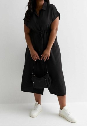 Zwarte jurk met knoopsluiting, korte mouwen, een getailleerde taille en een zwierige rok; gecombineerd met een kleine zwarte tas en witte sneakers.