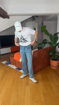 Młody mężczyzna poprawiający białą koszulkę z nadrukiem, ubrany w niebieskie dżinsy, sneakersy i białą czapkę, stojący w salonie z roślinami i meblami.