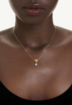 Goldene Kettenhalskette mit einem Knoten-Design und einem funkelnden Diamantakzent, präsentiert auf dunkler Haut und einem schwarzen Oberteil.