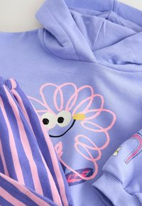 Felpa leggera color viola con una grafica di fiori giocosa, abbinata a pantaloni a righe viola e rosa, caratterizzati da un tessuto morbido e un design casual.