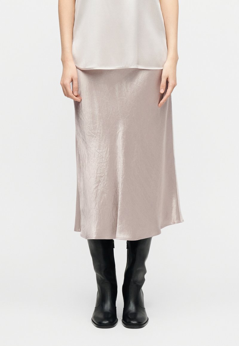 Κατώτερο μέρος σώματος ενός ατόμου που φορείει μια ανοιχτό μπεζ σατέν φούστα και μαύρες δερμάτινες μπότες έως το γόνατο, στέκεται μπροστά σε λευκό απλό φόντο.