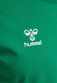 Grön bomullst-shirt med en vit Hummel-logotyp som innehåller en stiliserad bi och chevronmönster. Slät textur, rund halsringning.