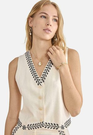 Joven mujer con cabello rubio lleva un top sin mangas color crema con bordado de hojas negras, joyas de perlas doradas y una pulsera dorada con forma de serpiente.