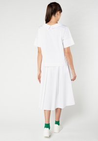 Vestito bianco con maniche corte e colletto, caratterizzato da una gonna plissettata. Tessuto liscio, design semplice, indossato con calze verdi e scarpe bianche.
