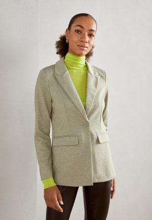 Blazer gris clair avec un bouton unique, revers en notch et deux poches avant, associé à un pull à col roulé vert vif et un pantalon marron.