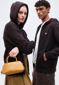 Γυναίκα με μαύρη φόρμα με κουκούλα που κρατά κίτρινη τσάντα, άνδρας με μαύρη φόρμα με κουκούλα και λευκή μπλούζα, και οι δύο κοιτάζουν μπροστά σε ομοιόμορφο φόντο.