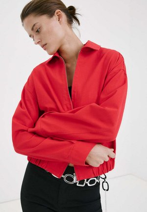 Młoda kobieta z włosami związanymi w kok, ubrana w jaskrawoczerwoną kurtkę na suwak i czarne spodnie z srebrnym łańcuchowym paskiem, pozuje z założonymi ramionami.