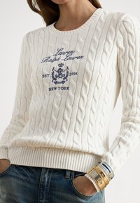 Beli pulover z vzorcem pletenja, z vezenim mornarsko modrim logotipom in datumom. Ima rebrast rob in manšete. Oblečen je z modrimi kavbojkami in kovinskimi zapestnicami.