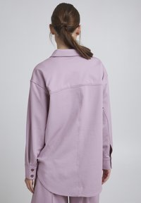 Oversized lavanda košulja od mekog materijala ima klasičan ovratnik, dugmad na manžetama i zakrivljen rub, s jednostavnim i urednim dizajnom.