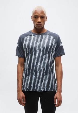 Jeune homme aux cheveux blonds courts portant un t-shirt de sport graphique à manches courtes noir et gris et un pantalon noir, debout devant un fond blanc.
