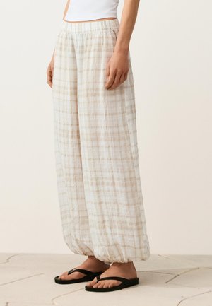 Žena v pohodlných béžových károvaných nohaviciach so zbieranými lemami, ktoré sú kombinované s čiernymi žabkami, stojaca na dlažobnej podlahe pred jednoduchým pozadím.