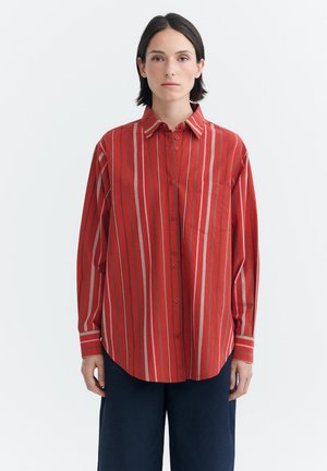 Femme portant une chemise rouge oversize à rayures verticales et un pantalon large foncé, debout devant un fond blanc uni.