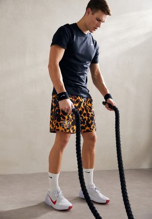 Hombre atlético con camiseta negra y pantalones cortos de estampado de leopardo sosteniendo cuerdas de batalla, preparándose para hacer ejercicio frente a una pared lisa.