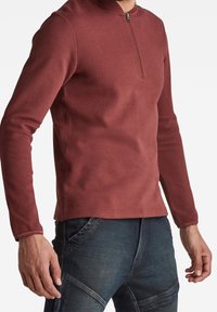 Haut à manches longues zippé en tissu rouge profond, avec un col montant, une coupe ajustée et une texture lisse.