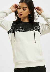 Sudadera con capucha en crema y negro, con una sección superior texturizada en negro con un patrón moteado y un bolsillo canguro en la parte frontal.