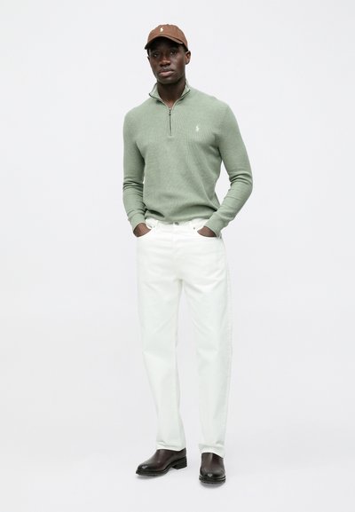 Világoszöld cipzáras pulóver, fehér farmernadrág, barna sapka, sötétbarna bokacsizma. Egyszerű kialakítás, kontrasztos színek és anyagok.