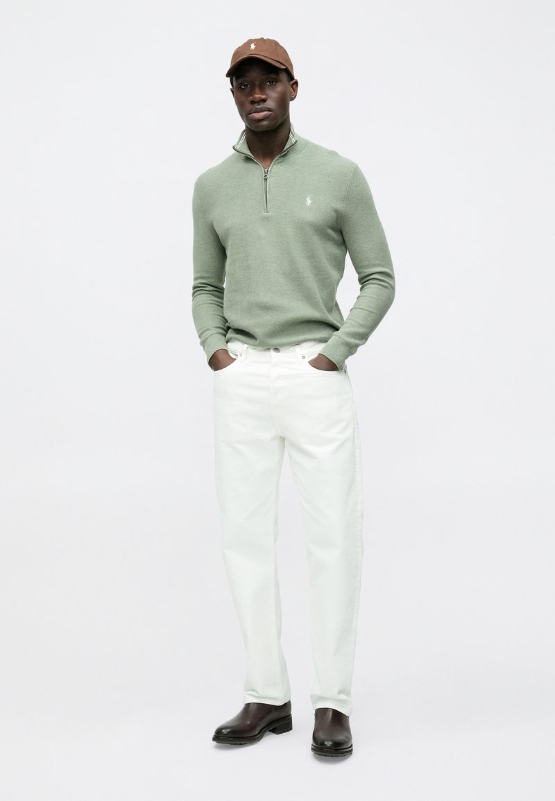 Maglione verde chiaro con cerniera, jeans bianchi, cappello marrone, stivaletti marrone scuro. Design semplice con colori e texture a contrasto.