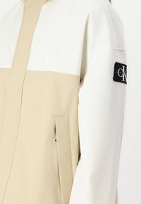 Jachetă crem și albă cu fermoar, cu un finisaj texturat, având un buzunar lateral și un patch cu logo-ul negru Calvin Klein pe mâneca stângă.