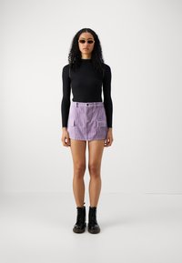 Čierny top s dlhým rukávom, levanduľová kordová sukňa s prednými vreckami a čierne šnurovacie topánky. Modelka nosí oválne slnečné okuliare.
