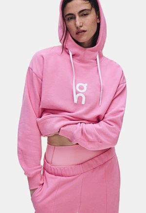 Femme portant un sweat à capuche rose avec des cordons blancs et un jogging rose assorti, soulevant légèrement le sweat pour révéler une ceinture en dessous.
