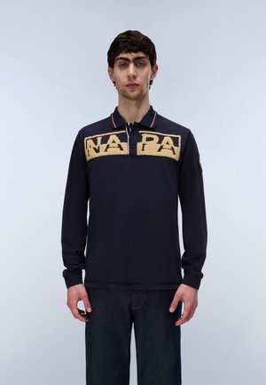 Joven con un polo azul marino de manga larga con las letras texturizadas en beige "NAPA" en el pecho y vaqueros azul oscuro.