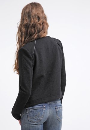 Černá textured bunda s cropped designem a čistými liniemi. Obsahuje kontrastní švy a kulatý výstřih. V kombinaci s modrými džíny.