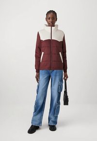 Moteris vilkinti bordo ir kremine pūkine striuke, mėlynomis krovinių džinsomis, juodais batais, laiko juodą rankinę, stovinti prieš vienodą foną.