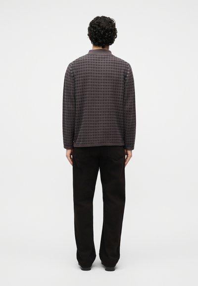 Vyras su garbanotais plaukais, dėvintis ilgomis rankovėmis margintą tamsiai pilką megztinį ir juodas kelnes, stovintis priešais paprastą šviesų foną.