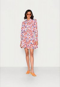Vestido floral com mangas longas, base rosa e padrões de folhas coloridas. Detalhes na barra e na gola em babados. Combinado com sandálias de salto laranja.