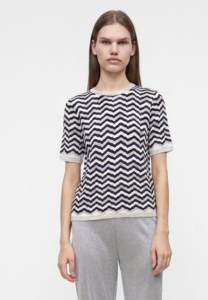Jeune personne aux cheveux raides portant une chemise à manches courtes à rayures ondulées noires et blanches et un pantalon gris clair, debout devant un fond uni.