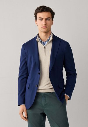 Young man wearing a navy blazer, beige quarter-zip sweater over a striped shirt, and dark green pants, standing against a plain background.