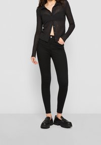 Kvinna iklädd genomskinlig svart långärmad skjorta med knappar, svarta höga skinnyjeans och svarta kraftiga loafers, stående mot vit bakgrund.