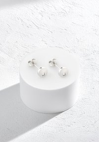 Silverfärgade stiftörhängen med en cirkulär skiva och en central fyrkantig accent, visade på en vit cirkulär bas mot en texturerad bakgrund.